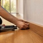 Mężczyzna mocuje drewnianą listwę przypodłogową przy ścianie za pomocą narzędzi 马会